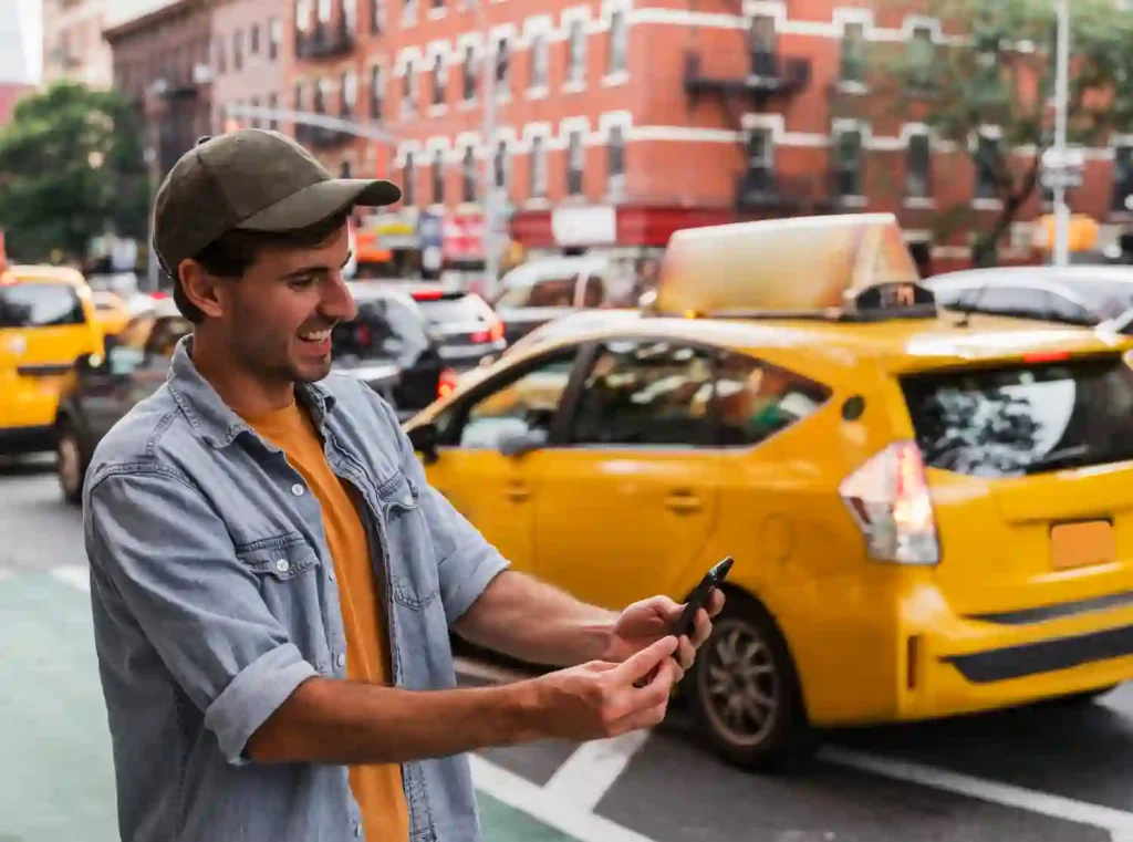 A smiling man in a cap takes a selfie on a busy city street with yellow taxis passing by; the scene is lively and urban, with a cheerful vibe. Romsey Cabs