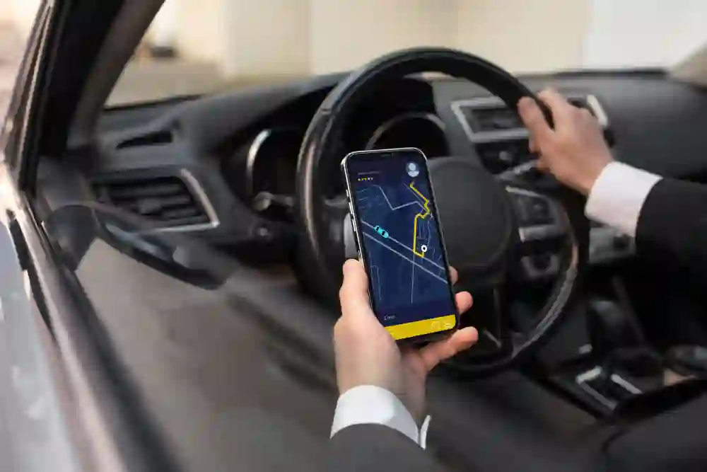 A person in a suit drives a car while holding a smartphone displaying a navigation app. The focus is on the app, suggesting a business trip or commute. Romsey Cabs